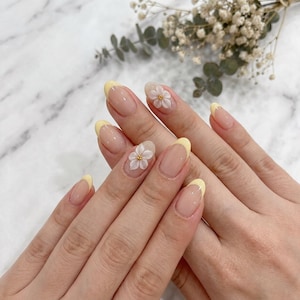 May include: Oval-shaped nails with a light pink base and yellow tips. Some nails have a 3D white flower with a yellow centre. The nails are on a hand, with a marble background.