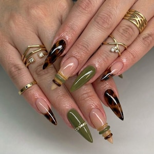 May include: A set of gold rings worn on fingers with long, pointed nails featuring a mix of solid olive green polish, tortoise shell patterns, and clear tips. Some nails are accented with thin gold bands or small embedded crystals.