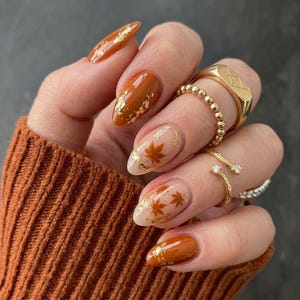 May include: A hand with almond-shaped nails featuring a mix of solid burnt orange polish and clear base coats adorned with gold leaf flakes and small brown maple leaf decals. The fingers are decorated with four gold rings of varying designs.