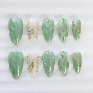 Op de afbeelding: Een set van tien kunstnagels in diverse ontwerpen. De nagels hebben een lichtgroen marmer effect, gouden vlinder accenten, bladsnijwerk en gouden lijnkunst. De nagels zijn ovaal van vorm en zijn gerangschikt op een witte ondergrond.