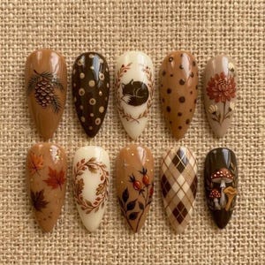 May include: A set of ten almond-shaped press-on nails featuring autumn-themed designs including a pinecone, brown polka dots, a sleeping fox, a floral bloom, maple leaves, an oak leaf wreath, rose hips, a brown plaid pattern, and mushrooms.
