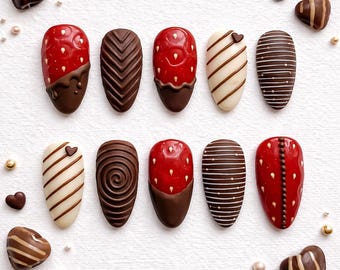 May include: A collection of ten artificial nails with various designs. Some nails are red with white dots and dipped in chocolate. Others feature chocolate patterns, including stripes, chevrons, and swirls. Decorative chocolate hearts and pearls are scattered around the nails.