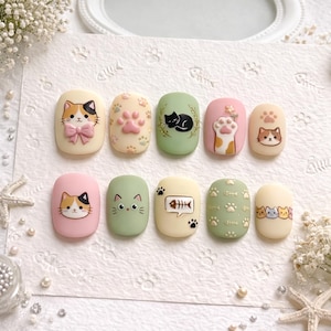 Puede incluir: Un conjunto de diez uñas postizas con diseños de gatos. Las uñas presentan varios colores, incluyendo rosa, verde y crema, con caras de gato, huellas de patas y gráficos de espinas de pescado. Algunas uñas tienen elementos 3D.