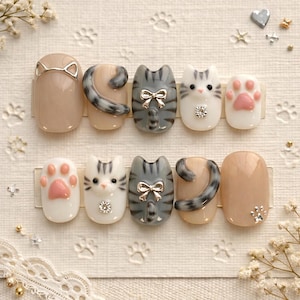 Puede incluir: Un conjunto de diez uñas postizas con temática de gatos. Los diseños incluyen caras de gato, huellas de patas y colas de gato. Los colores incluyen beige, gris, blanco y rosa. Algunas uñas presentan lazos plateados y pedrería. Las uñas están dispuestas sobre una superficie blanca.