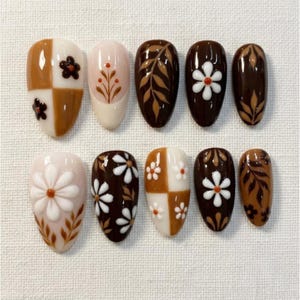 May include: A set of ten press-on nails featuring a brown, cream, and white color palette. The designs include checkered patterns, raised white floral accents, and intricate brown leaf illustrations painted on almond-shaped nail tips.