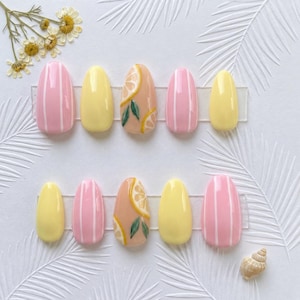 May include: A set of press-on nails with a summer theme. The nails feature a variety of designs, including solid yellow, pink with white stripes, and a nail with a lemon slice design. The nails are oval-shaped and arranged on a clear plastic sheet.