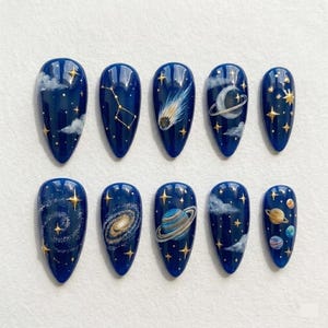 May include: A set of ten almond-shaped press-on nails featuring a deep navy blue base with hand-painted celestial designs including gold stars, white clouds, a comet, a crescent moon with a ring, a spiral galaxy, and various colorful planets.