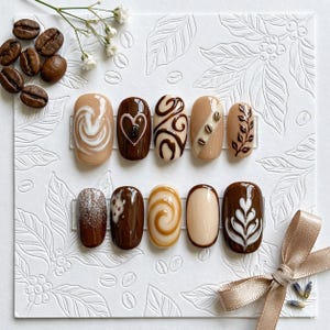 May include: A set of ten press-on nails featuring coffee-themed designs in shades of brown, cream, and tan. The nails display patterns including swirls, hearts, coffee beans, leaf motifs, and latte art, arranged in two rows on a white textured card.
