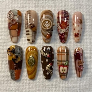 Puede incluir: Un conjunto de diez uñas postizas con diseños de temática otoñal que incluyen patrones de cuadros, arte de café, bellotas, hojas, botones, orugas, manzanas y estrellas en tonos marrón, naranja, crema y burdeos con acabados brillantes.