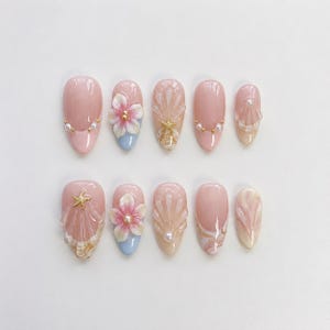 Könnte beinhalten: Ein Set aus zehn künstlichen Nägeln in hellem Rosa, verziert mit verschiedenen Designs. Einige Nägel weisen 3D-Blütenakzente auf, während andere Muschel- und Seesternverzierungen zeigen. Perlenakzente verleihen der Nagelkunst Eleganz.