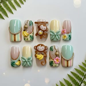 Könnte beinhalten: Ein Set aus zehn künstlichen Nägeln mit tropischem Thema. Designs beinhalten einen Affen mit Bananen, Blumen und grünen Blättern. Farben sind Hellgrün, Gelb, Rosa und Braun. Einige Nägel haben einen seilartigen Rand.