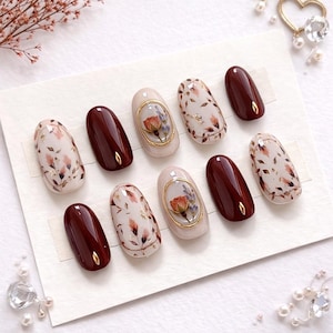 May include: A set of ten press-on nails featuring a mix of glossy deep burgundy nails with small gold accents and cream-colored nails decorated with delicate floral patterns, dried flower inclusions, and gold-toned oval frames.