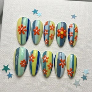 May include: A set of ten almond-shaped press-on nails featuring vertical stripes in shades of light blue, yellow, and dark blue. Each nail is decorated with raised, three-dimensional orange flower appliqués with small yellow centers.