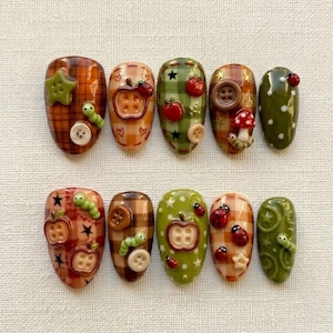 Op de afbeelding: Een set van tien kunstnagels met diverse ontwerpen, waaronder bruine en groene ruitpatronen, olijfgroene en crèmekleurige achtergronden. De versieringen bestaan uit 3D-knopen, appeltjes, lieveheersbeestjes, sterren, paddenstoelen en rupsen.