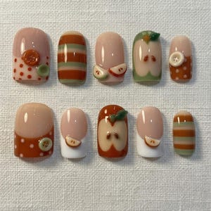 May include: A set of ten press-on nails featuring a variety of designs including orange and green stripes, polka dots with small attached buttons, and raised 3D apple slice motifs on a glossy nude, white, and orange base.