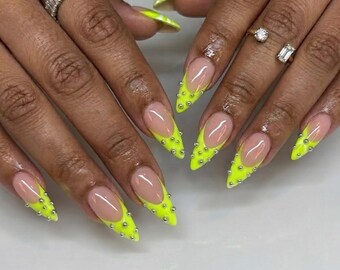 Op de afbeelding: Puntige, amandelvormige nagels in een French manicure-stijl met levendige neongele uiteinden. Elk neongeel uiteinde is versierd met kleine, verhoogde zilveren metalen studs die in een stippenpatroon over het gekleurde gedeelte zijn geplaatst.