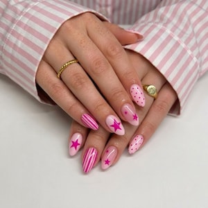Pode incluir: Unhas de formato oval com uma base rosa claro e designs de nail art em magenta vibrante, incluindo listras verticais, pequenas bolinhas e estrelas de cinco pontas. Dois anéis de metal dourado são usados nos dedos das mãos.