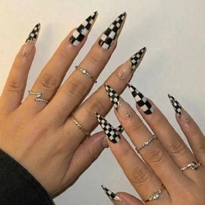 Könnte beinhalten: Lange, stiletto-förmige künstliche Fingernägel mit schwarz-weißem Schachbrettmuster an den Spitzen, verziert mit kleinen goldfarbenen Nieten an der Nagelbasis, kombiniert mit mehreren dünnen goldfarbenen Metallringen an den Fingern.