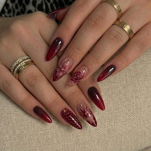 May include: Almond-shaped nails featuring a deep burgundy ombre gradient, intricate hand-painted floral motifs, and a textured honeycomb pattern. The design is accented with small gold studs and paired with three gold bands worn on the fingers.