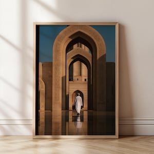 May include: Framed print of a mosque interior. A person in white robes walks through arched doorways. The architecture is beige with intricate carvings. The sky is a deep blue. The frame is light brown.