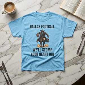 May include: Light blue t-shirt with the text "DALLAS FOOTBALL WE'LL STOMP YOUR HEART OUT". The shirt features a graphic of a cowboy-like figure holding a football. The shirt is on a marble surface.