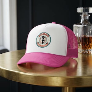 May include: A pink and white Zumba trucker hat with a curved brim. The hat features a circular emblem with the word "Zumba" and a dancing figure. The mesh back is pink, and the hat sits on a gold surface.