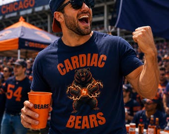 May include: A navy blue t-shirt featuring an orange graphic of a bear surrounded by lightning bolts with the text CARDIAC BEARS in orange. The shirt is worn with a navy blue baseball cap and sunglasses, while holding an orange plastic cup.