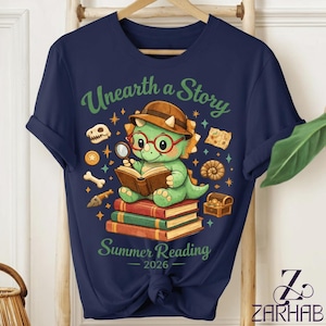 May include: Navy blue t-shirt featuring a green dinosaur wearing glasses and a hat, reading a book. The shirt has the text "Unearth a Story Summer Reading 2026" with illustrations of fossils and a treasure chest.