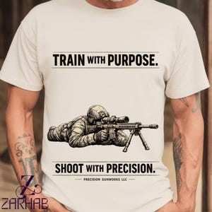 以下が含まれることがあります： クリーム色のTシャツ。スコープ付きのライフルを構える兵士の詳細なイラストが描かれています。「TRAIN WITH PURPOSE.」の文字がイラストの上に、「SHOOT WITH PRECISION.」の文字が下にあります。シャツには「PRECISION GUNWORKS LLC」の文字も含まれています。