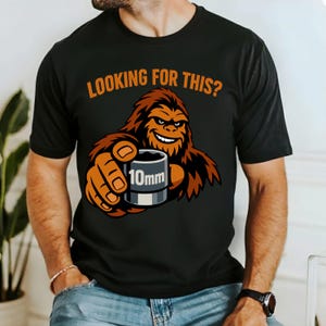 Könnte beinhalten: Schwarzes T-Shirt mit dem Text "LOOKING FOR THIS?" in Orange. Das Shirt zeigt eine Cartoon-Figur von Bigfoot, die einen 10-mm-Steckschlüssel hält. Der Bigfoot ist braun und orange, mit einem lächelnden Gesichtsausdruck.