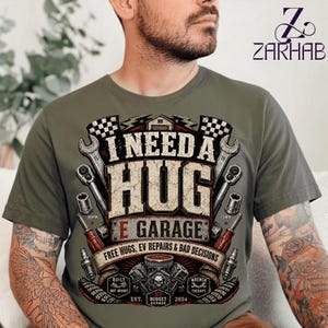 Puede incluir: Una camiseta verde oliva con un estampado gráfico que incluye el texto I NEED A HUG E GARAGE FREE HUGS, EV REPAIRS & BAD DECISIONS. El diseño presenta ilustraciones de llaves inglesas, vasos, bujías y un bloque de motor con banderas a cuadros.