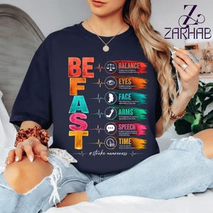 Puede incluir: Camiseta azul marino con el texto "BE FAST" en letras de colores. La camiseta presenta un gráfico con las palabras "BALANCE, EYES, FACE, ARMS, SPEECH, TIME" y el hashtag "#stroke awareness".