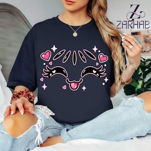 Könnte beinhalten: Ein dunkelblaues Kurzarm-T-Shirt mit einem Grafikdruck auf der Vorderseite, der ein stilisiertes Gesicht mit geschlossenen Augen, langen Wimpern, einer herausgestreckten Zunge sowie rosa Herzen und weißen vierzackigen Sternen zeigt.