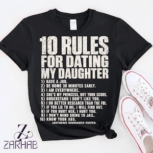 Puede incluir: Una camiseta negra de manga corta con texto blanco desgastado que dice 10 RULES FOR DATING MY DAUGHTER seguido de una lista numerada de diez reglas y las palabras ABSTINENCE BOUNDARIES-CURFEW impresas en una fuente sans-serif negrita.