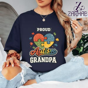 Puede incluir: Camiseta azul marino con un diseño de rompecabezas en forma de corazón en rojo, naranja, verde y azul, con las palabras "PROUD Autism GRANDPA" en un estilo vintage. Prenda de vestir informal.