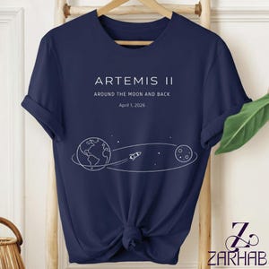 May include: Navy blue t-shirt with the text "ARTEMIS II" and "AROUND THE MOON AND BACK" with a graphic of Earth, a rocket, and the moon. The date "April 1, 2026" is also printed on the shirt.