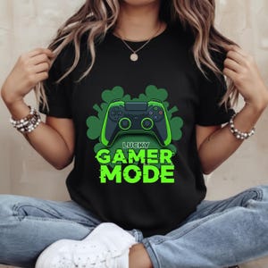 Puede incluir: Camiseta negra con un diseño de mando de videojuegos verde y negro y el texto "LUCKY GAMER MODE" en verde. El diseño incluye formas de trébol. La camiseta está hecha de un material suave.