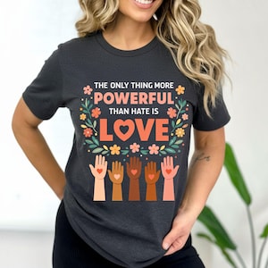 Puede incluir: Camiseta gris oscuro con el texto "THE ONLY THING MORE POWERFUL THAN HATE IS LOVE." El diseño incluye una corona floral, un corazón y manos en varios tonos de piel. La camiseta es de manga corta.