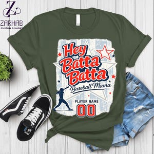 May include: Olive green t-shirt with a baseball-themed graphic. The design features the text "Hey Batta Batta Baseball Mama" in red and white, along with a baseball player silhouette and the number "00". Black and white sneakers and denim shorts are also visible.