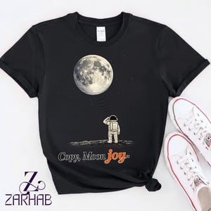 Puede incluir: Camiseta negra con un gráfico de un astronauta mirando la luna. El texto "Copy, Moon joy" está impreso debajo en naranja y blanco. Se ven zapatillas blancas con ribetes rojos.