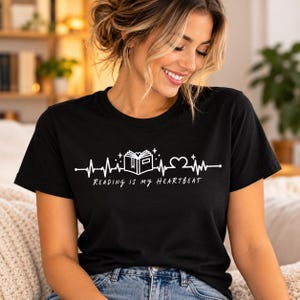 Puede incluir: Una camiseta negra de manga corta con un gráfico blanco de una línea de latido de corazón, un libro abierto y un corazón. El texto Reading is my heartbeat está impreso en blanco debajo del diseño gráfico en la parte frontal.