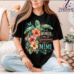 May include: A black t-shirt featuring a colorful graphic of a sea turtle surrounded by tropical hibiscus flowers and butterflies. The text reads, In a World Full of Grandmas Be a MIMI, with decorative flourishes and a small heart icon integrated into the design.