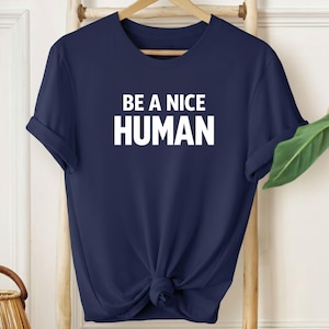 Puede incluir: Camiseta azul marino con el texto blanco "BE A NICE HUMAN". La camiseta está anudada en la parte inferior y cuelga de una percha de madera. El fondo incluye una escalera de madera y una planta verde.