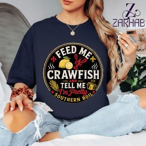 Puede incluir: Camiseta azul marino con un diseño circular que dice "FEED ME CRAWFISH AND TELL ME I'M PRETTY SOUTHERN BOIL". El diseño incluye un cangrejo de río, un limón, maíz y una taza de café. La camiseta tiene cuello redondo.