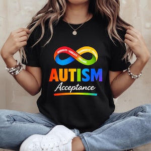 Puede incluir: Camiseta negra con un símbolo de infinito colorido y las palabras "ADVOCATE - EDUCATE - ACCOMMODATE - INCLUDE". Debajo, la palabra "AUTISM" está en colores del arcoíris, con "Acceptance" debajo. Una franja de arcoíris en la parte inferior.
