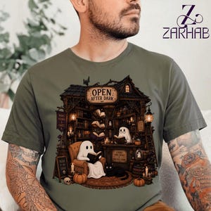 Puede incluir: Una camiseta verde oliva con un gráfico de un acogedor interior de librería oscura con el texto "OPEN AFTER DARK". La ilustración incluye dos fantasmas blancos leyendo libros, un gato negro, estanterías llenas de libros, velas y linternas.