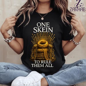 Puede incluir: Una camiseta negra de manga corta con un gráfico de un ovillo de lana dorado sobre un trono hecho de agujas de tejer. El texto dice ONE SKEIN TO RULE THEM ALL. El diseño está centrado, con el logotipo de ZARHAB en la esquina superior derecha.