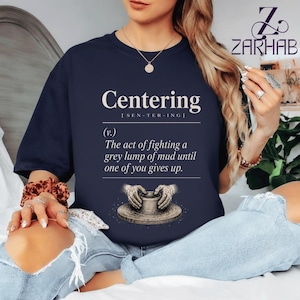 Puede incluir: Camiseta azul marino con la palabra "Centering" y su pronunciación entre corchetes. Debajo, una definición dice: "El acto de luchar contra un montón de barro gris hasta que uno de vosotros se rinda". Un gráfico de manos moldeando arcilla está debajo.