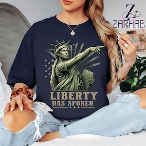 Puede incluir: Una camiseta azul marino con un gráfico de la Estatua de la Libertad señalando hacia adelante, sosteniendo una tableta, con un patrón estilizado de rayos de sol y estrellas de fondo. El texto "LIBERTY HAS SPOKEN" está impreso en oro debajo.