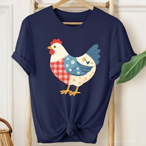 Puede incluir: Camiseta azul marino con un diseño caprichoso de gallina. La gallina tiene un patrón de cuadros rojos y blancos, detalles florales azules y un ala azul. Una ilustración colorida y juguetona.